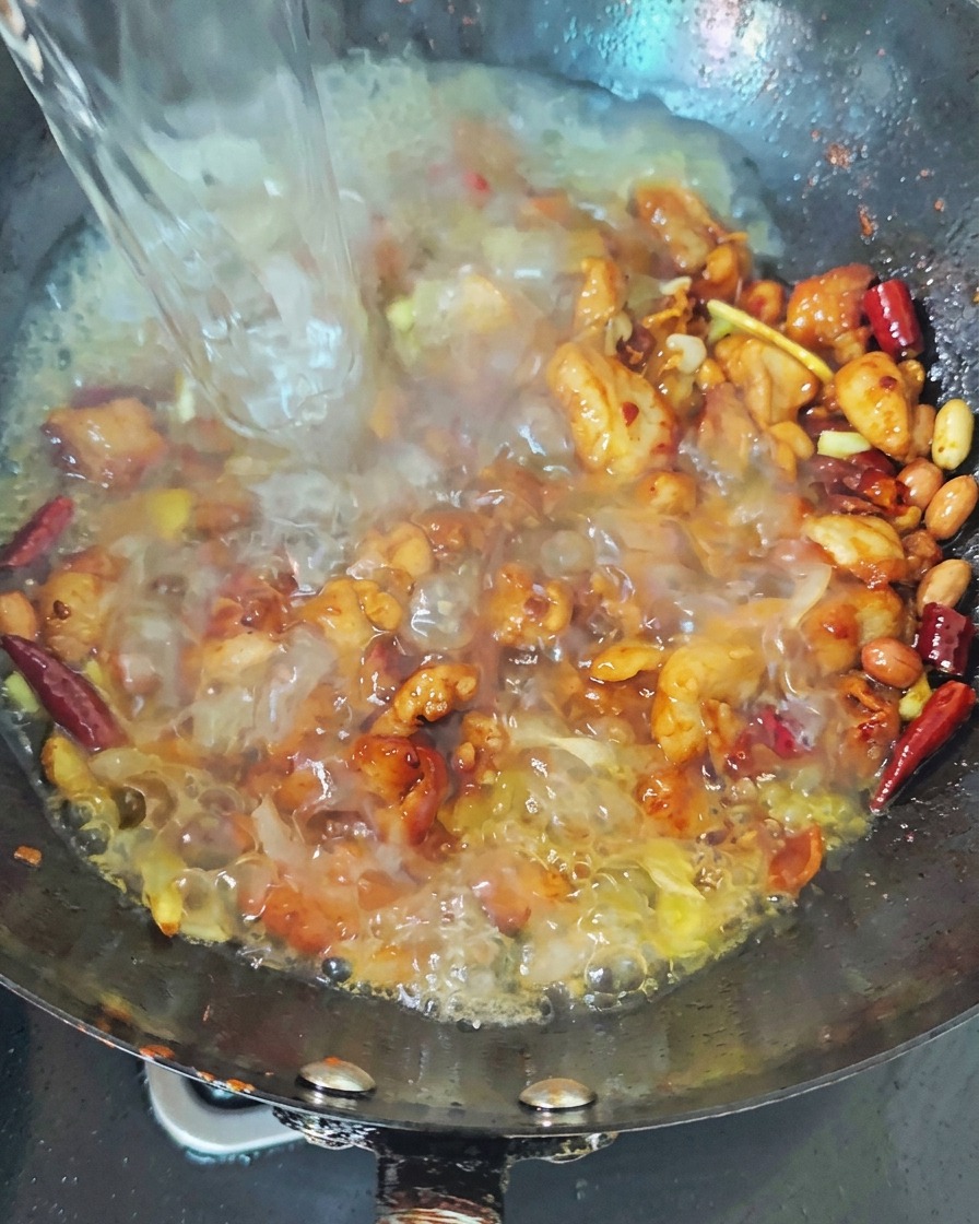 Chorro de agua hirviendo vertiéndose en un wok oscuro que contiene pollo sellado y chiles rojos, creando un caldo burbujeante.