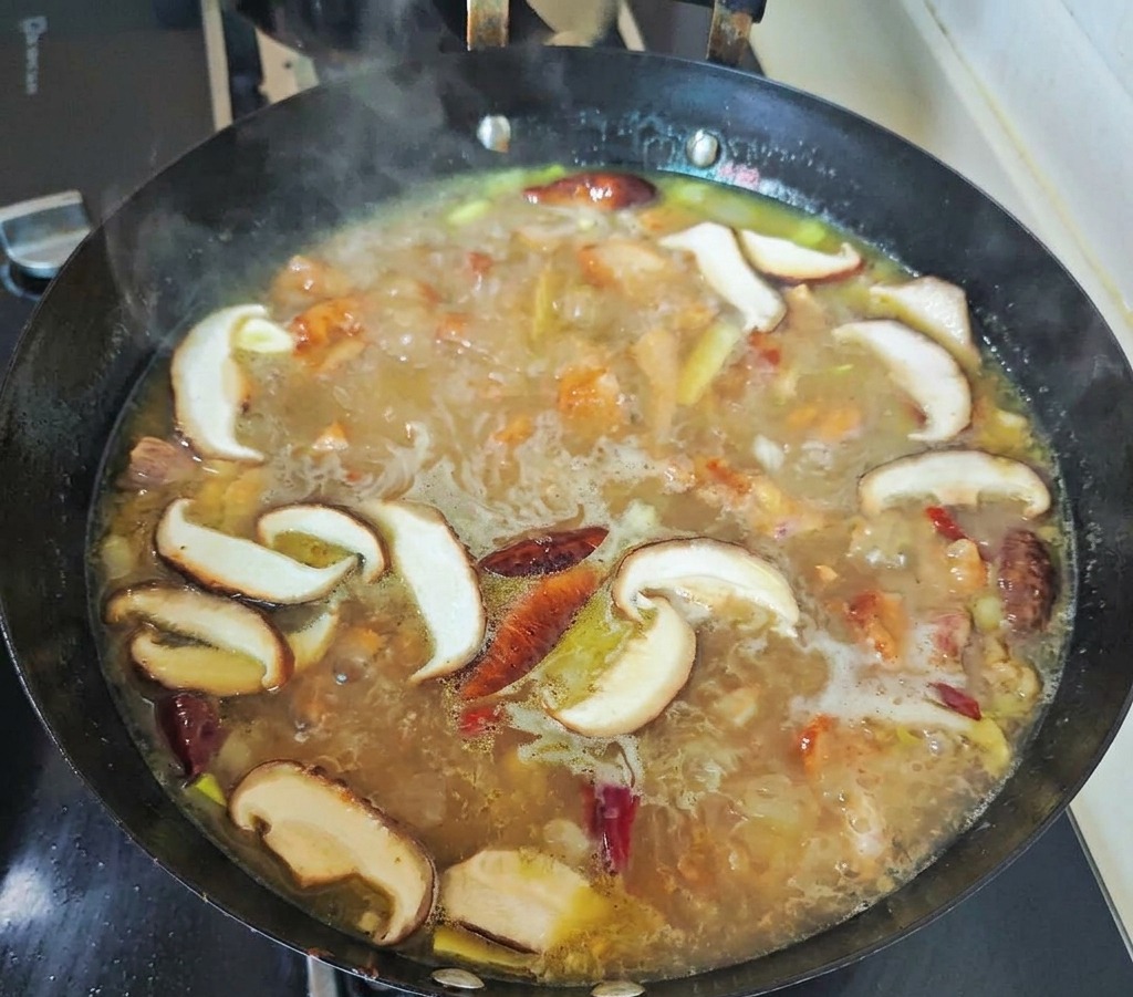 Setas shiitake en rodajas y chiles rojos hirviendo a fuego lento en un caldo de pollo dorado y burbujeante dentro de un wok negro.