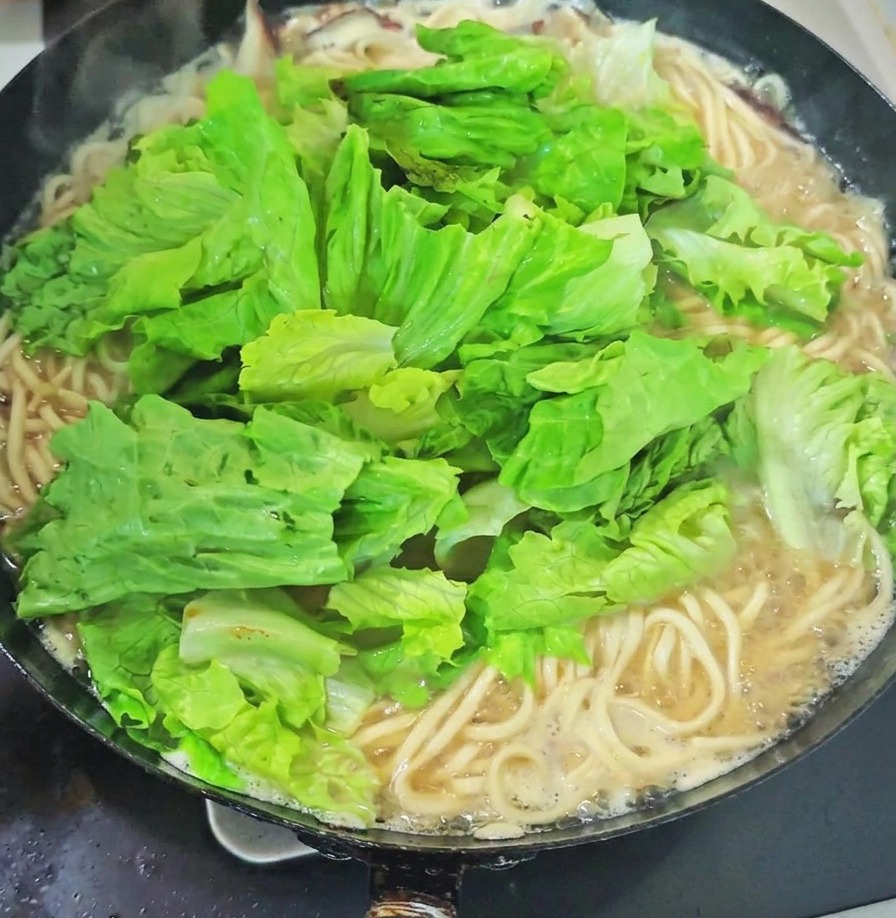 Un buen puñado de verduras de hoja verde fresca y brotes de ajo sobre los fideos en un wok de sopa hirviendo.