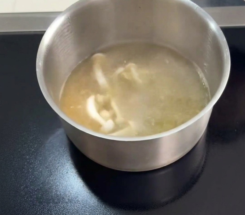 Setas de jade blanco y finas hebras naranjas de flores de cordyceps siendo añadidas a una olla con caldo claro de pollo hirviendo.