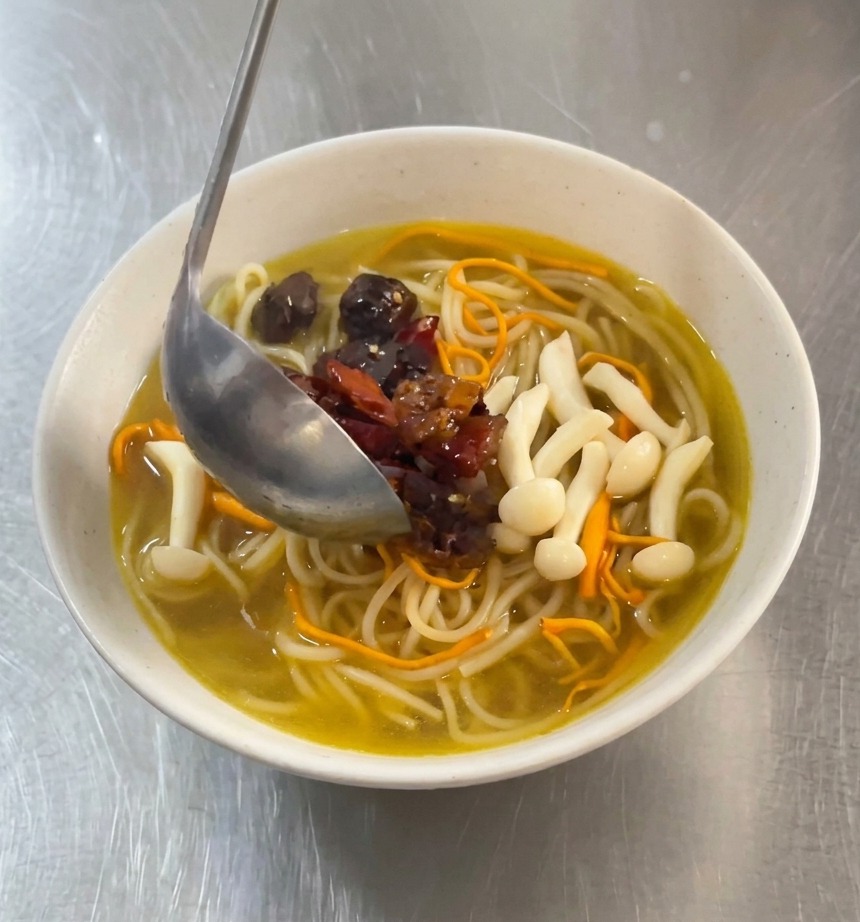 Un cucharón metálico sirviendo trozos de pollo picante marinado en rojo y chiles secos sobre un tazón de fideos blancos en caldo dorado con setas.