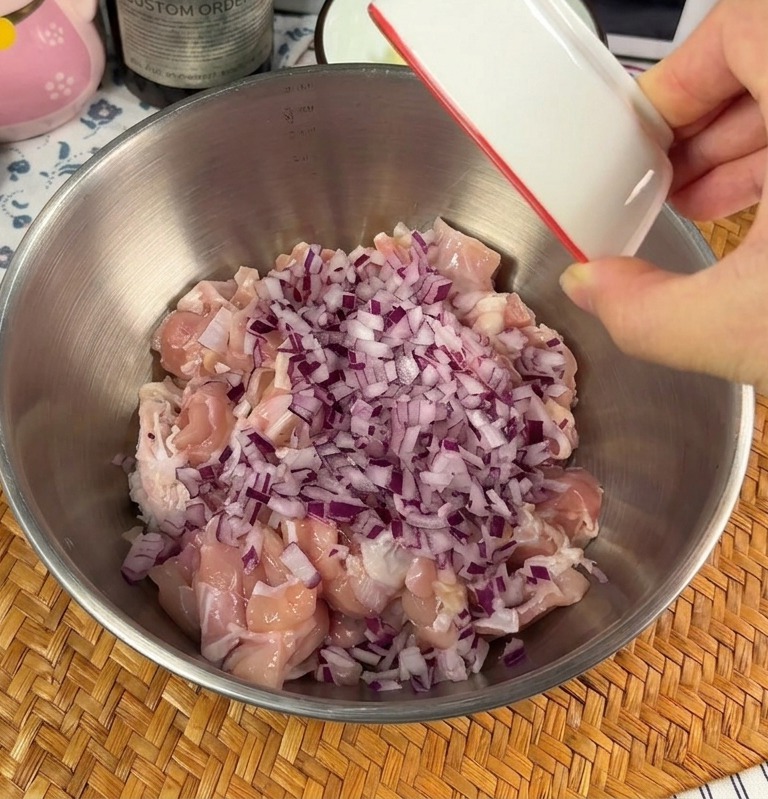 Una mano vertiendo un plato pequeño con cebolla morada picada y ajo picado sobre trozos de pollo crudo en un bol metálico.