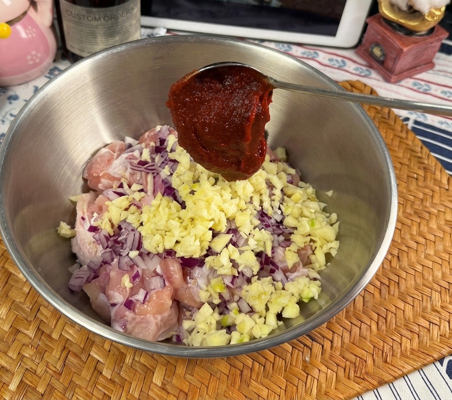 Una cuchara metálica con una porción generosa de pasta Gochujang de color rojo brillante sobre un bol con pollo crudo, ajo y cebolla.