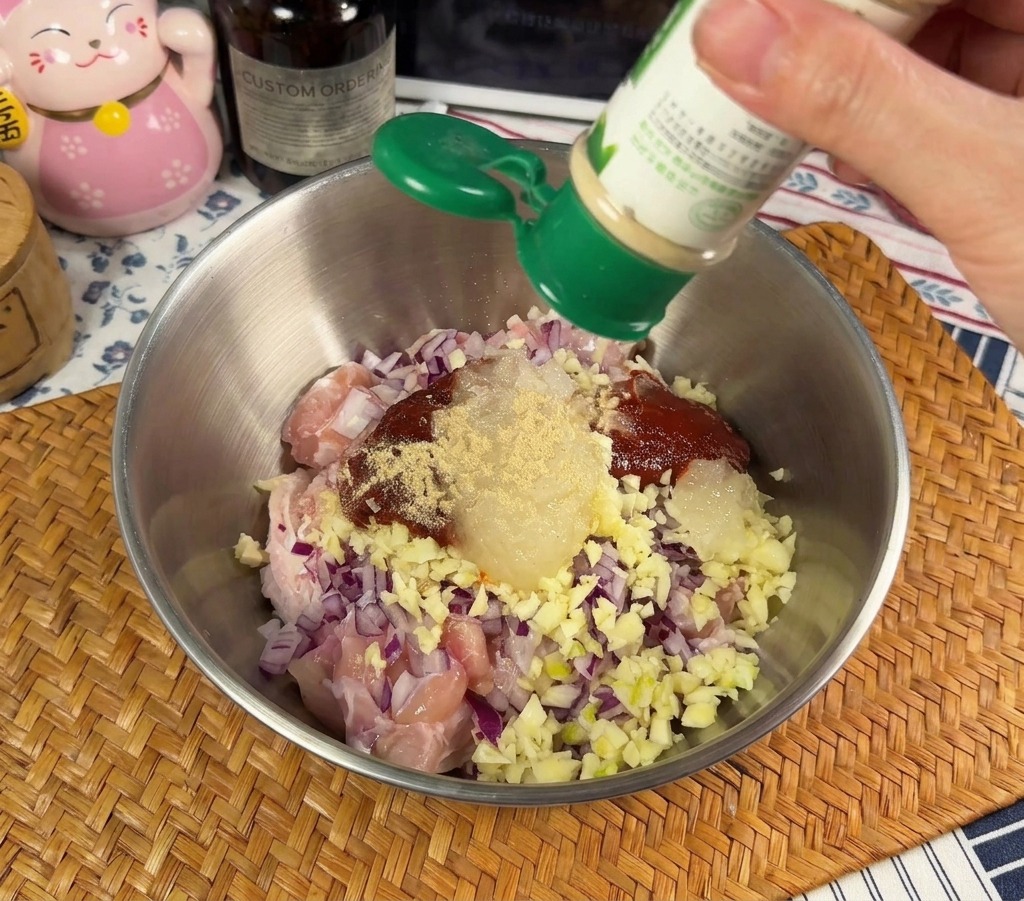 Una persona espolvoreando pimienta blanca en polvo desde un frasco verde en un bol de metal con pollo crudo y cebolla morada picada.