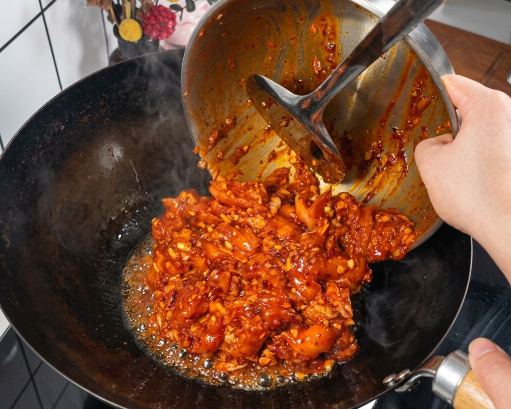 Trozos de pollo marinado en una salsa roja espesa cayendo desde un bol metálico hacia un wok negro caliente y humeante.