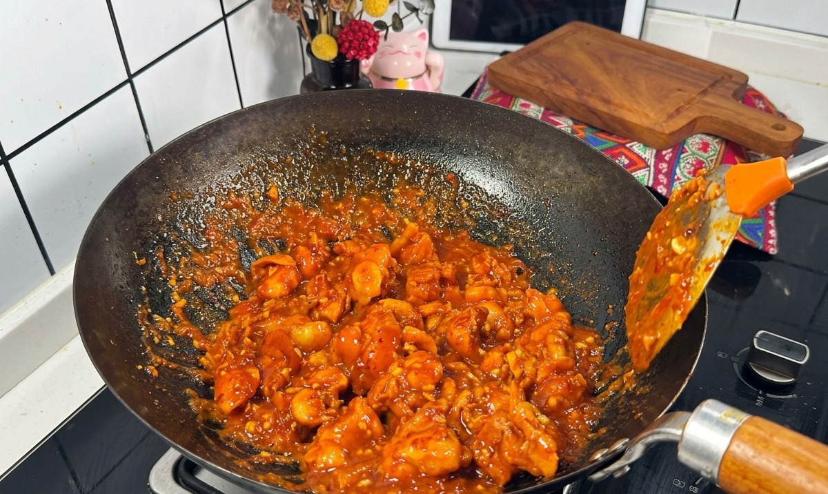 Trozos de pollo cubiertos con una salsa roja espesa y burbujeante siendo salteados en un wok negro con una espátula metálica.