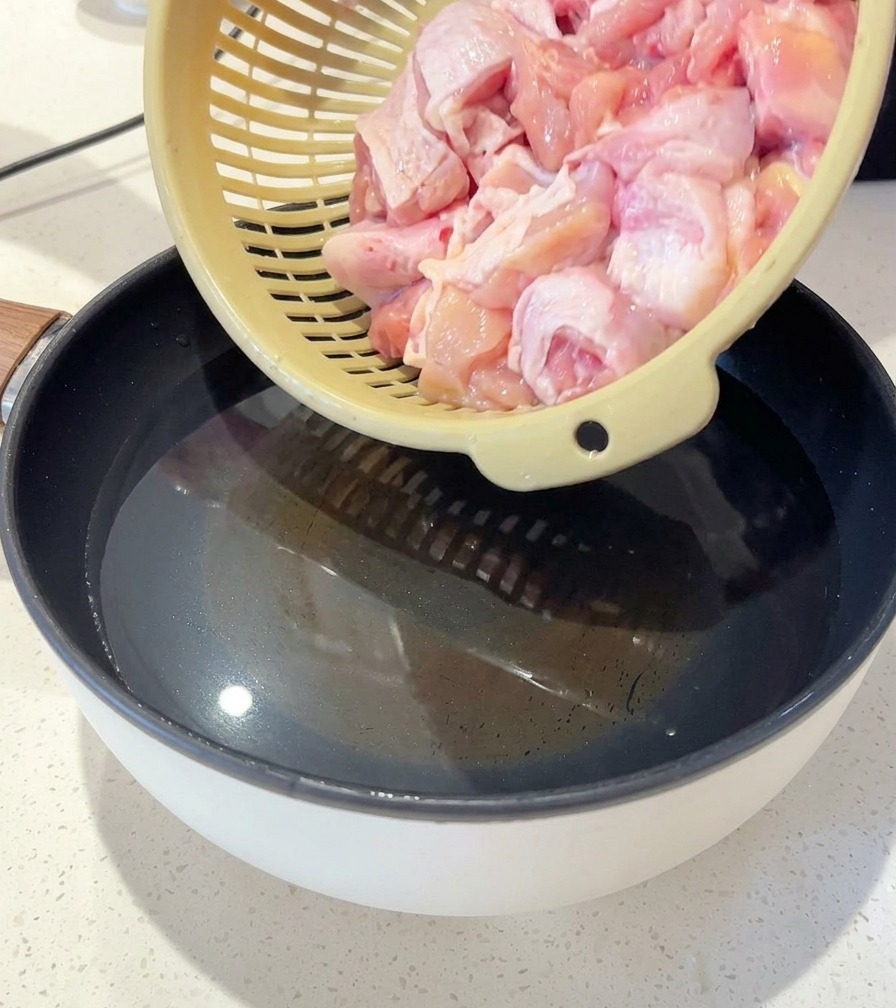 Una persona pasando trozos de pollo crudo desde un colador a una olla con agua, rodajas de jengibre y cebolleta.