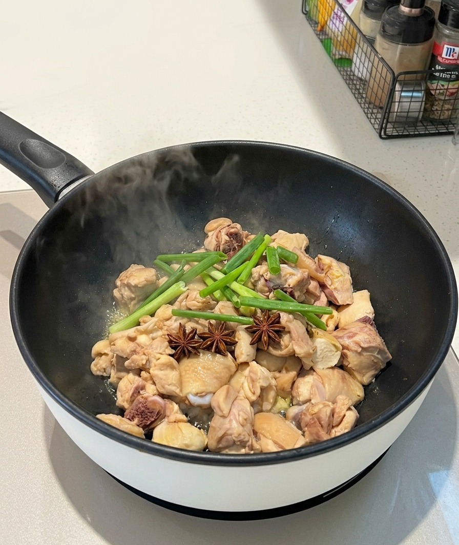 Anís estrellado entero y trozos de cebolleta verde colocados sobre los trozos de pollo en una sartén humeante.