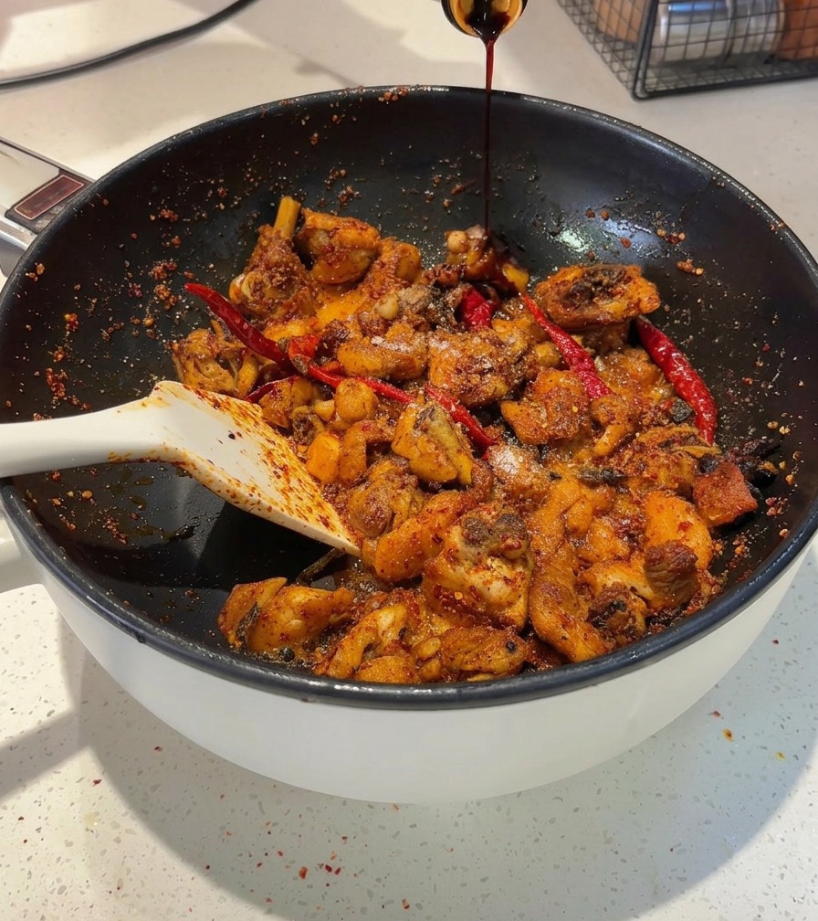 Salsa de soja oscura vertiéndose desde una botella en una sartén que contiene trozos de pollo picante.