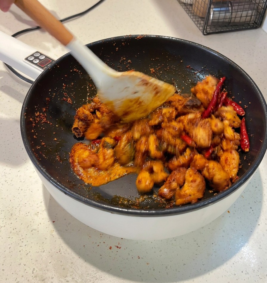 Una espátula blanca mezclando trozos de pollo en una salsa roja oscura dentro de una sartén hasta que cada trozo quede cubierto.