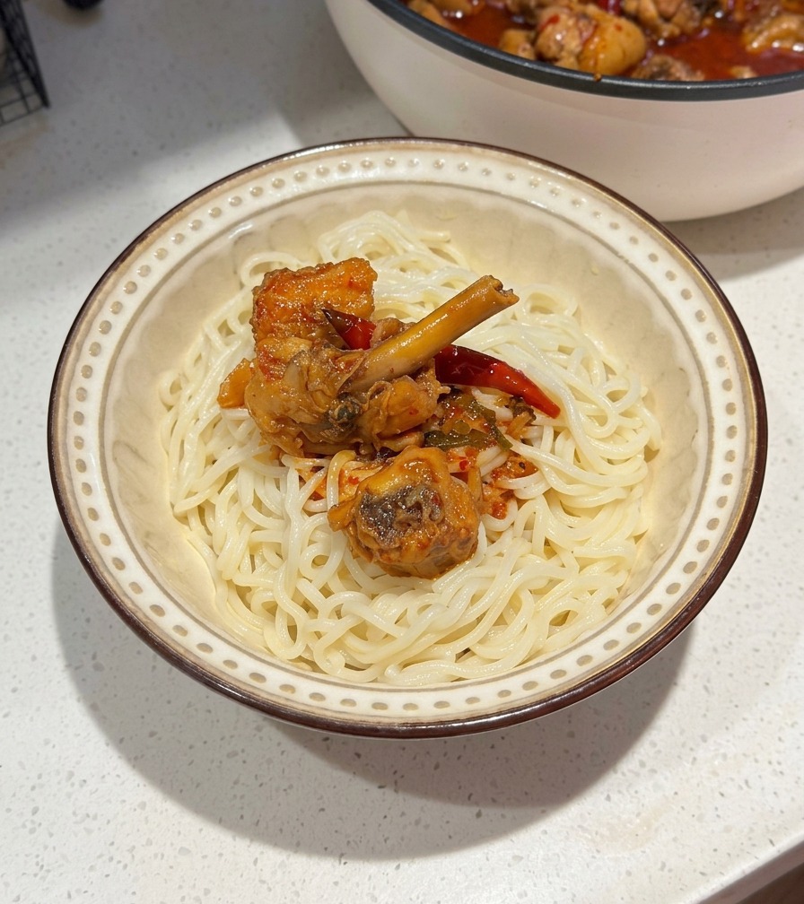 Un tazón de cerámica con fideos blancos cubiertos con pollo picante y salsa de chile rojo, con la sartén de cocción visible al fondo.