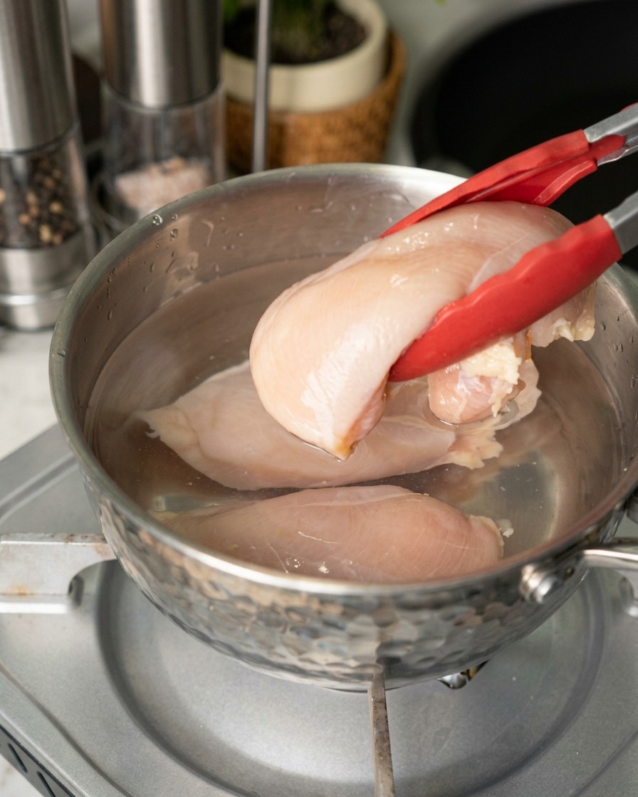 Una persona usando pinzas con punta roja para colocar pechugas de pollo crudas en una olla de acero inoxidable con agua.