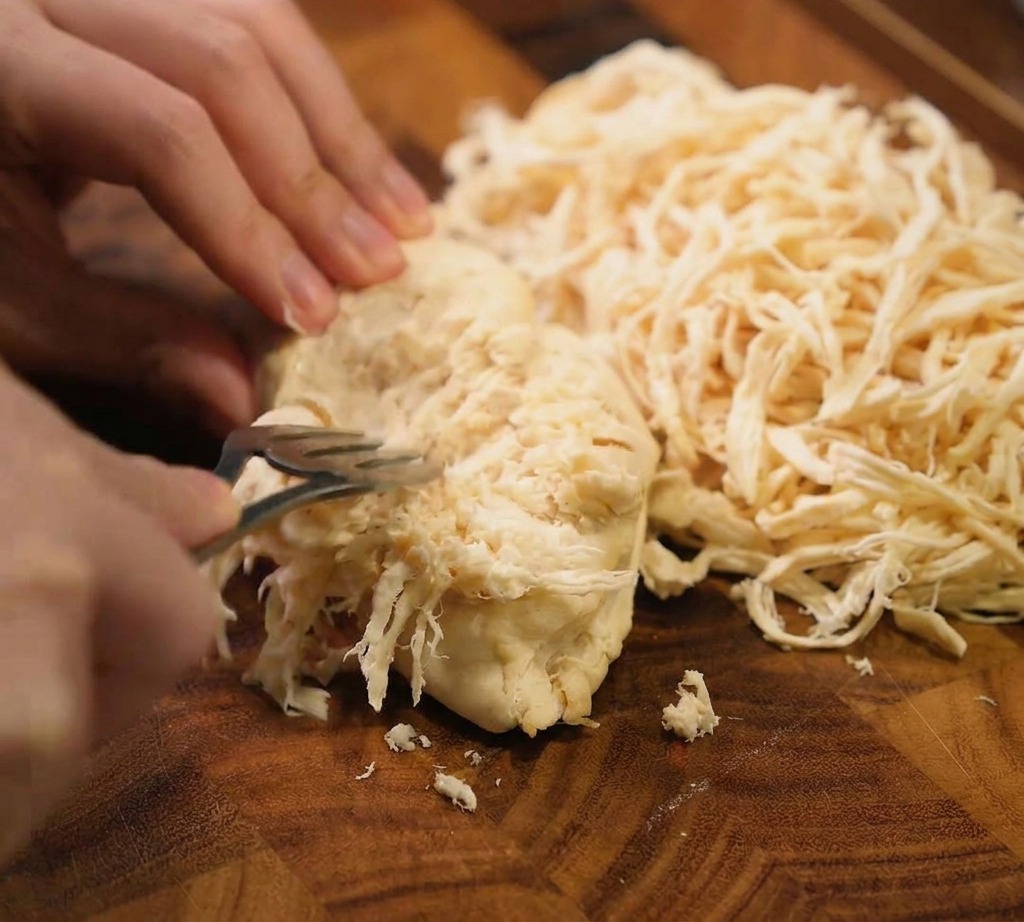 Un par de tenedores de metal desmechando una pechuga de pollo cocida en tiras finas sobre una tabla de cortar de madera oscura.