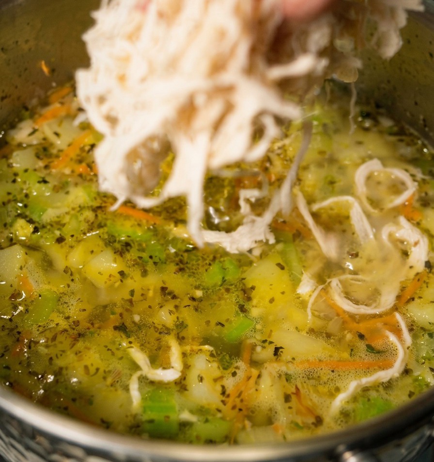 Una mano echando carne de pollo blanca desmechada en una olla de sopa de verduras a fuego lento con zanahorias y apio visibles.