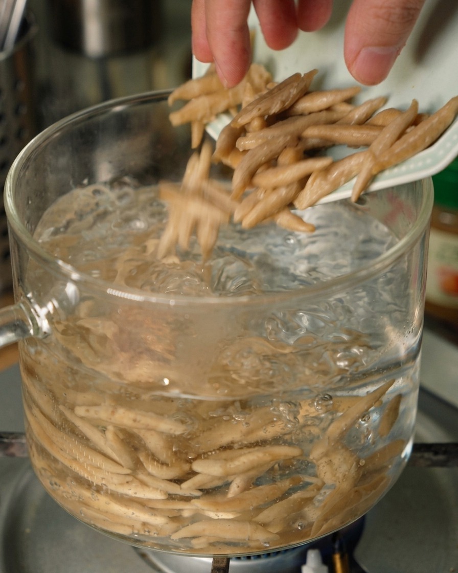 Fideos de avena afilados cayendo desde un plato pequeño en una olla de vidrio transparente con agua hirviendo a borbotones.