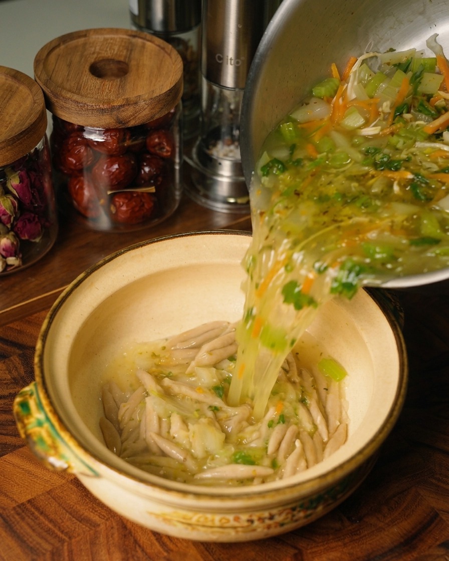 Sopa caliente de pollo y verduras vertiéndose desde una olla de acero inoxidable en un tazón de cerámica que contiene fideos de avena cocidos.