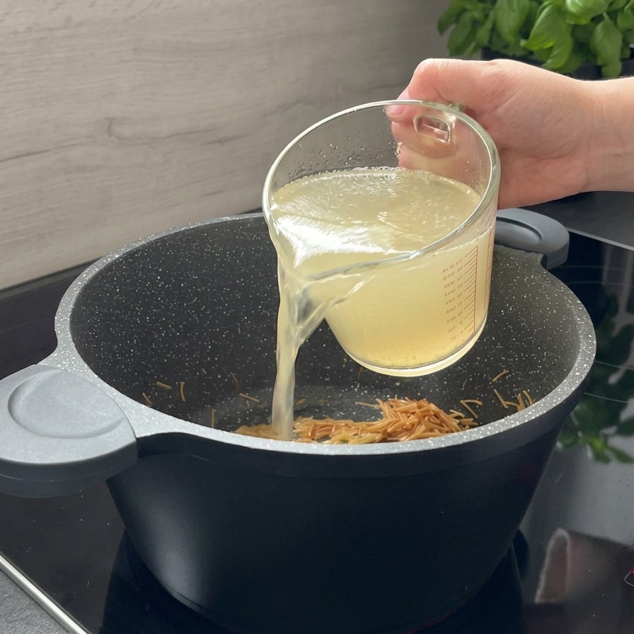 Caldo de pollo limpio vertiéndose desde una jarra medidora de cristal a una olla negra con pasta tostada.