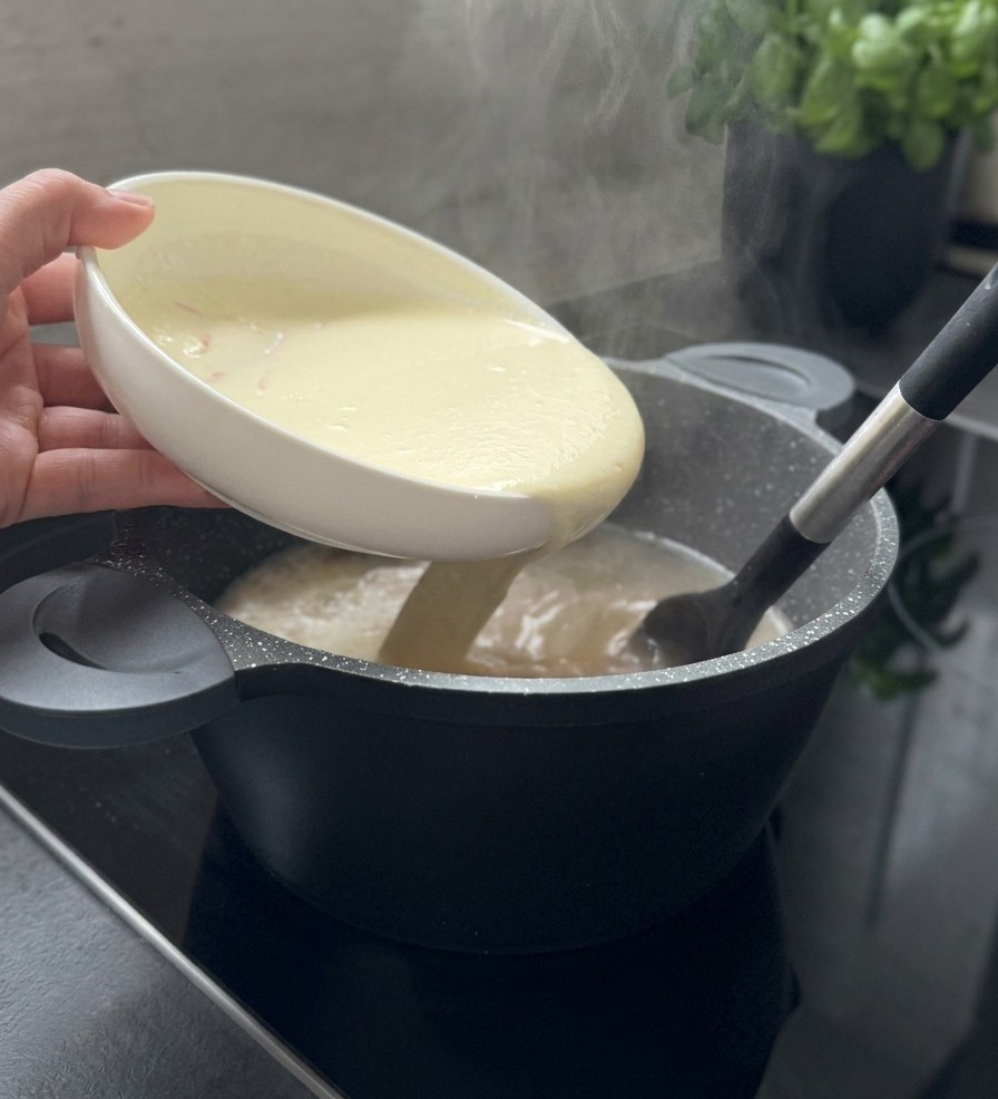 La crema blanca de yogur vertiéndose desde un bol en una olla de sopa humeante mientras se remueve con una espátula negra.