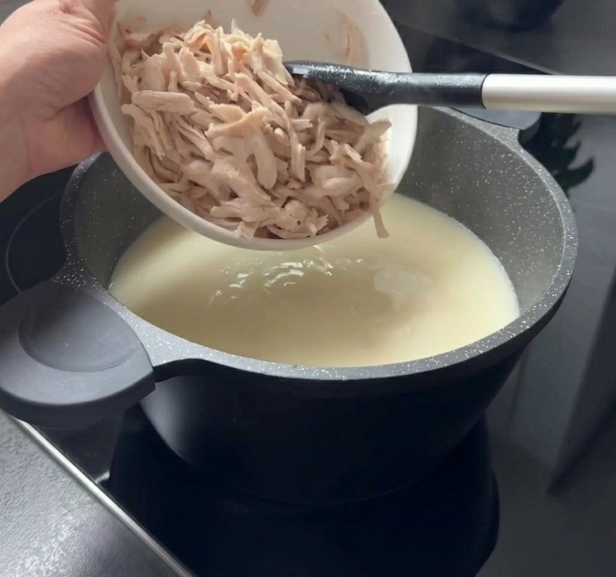 Trozos de pollo cocido y deshilachado cayendo desde un bol blanco a una olla de sopa blanca y cremosa.