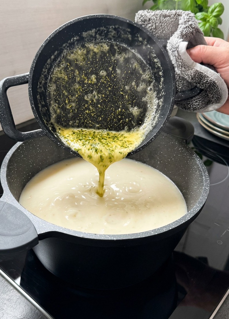 Mantequilla amarilla derretida con hojas de menta seca vertiéndose desde un cazo negro a una sopa cremosa blanca.