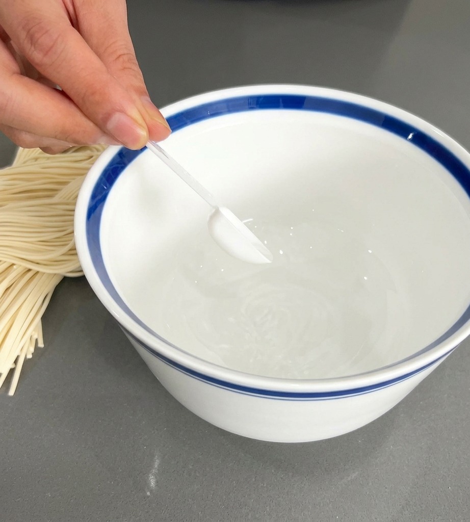 Una mano que sostiene una pequeña cuchara transparente y coloca sazonador blanco en un cuenco blanco con borde azul.