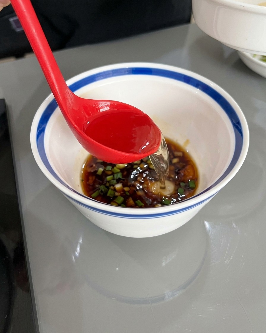 Un cazo rojo vertiendo agua caliente en el cuenco con la salsa de soja y los aromáticos picados.