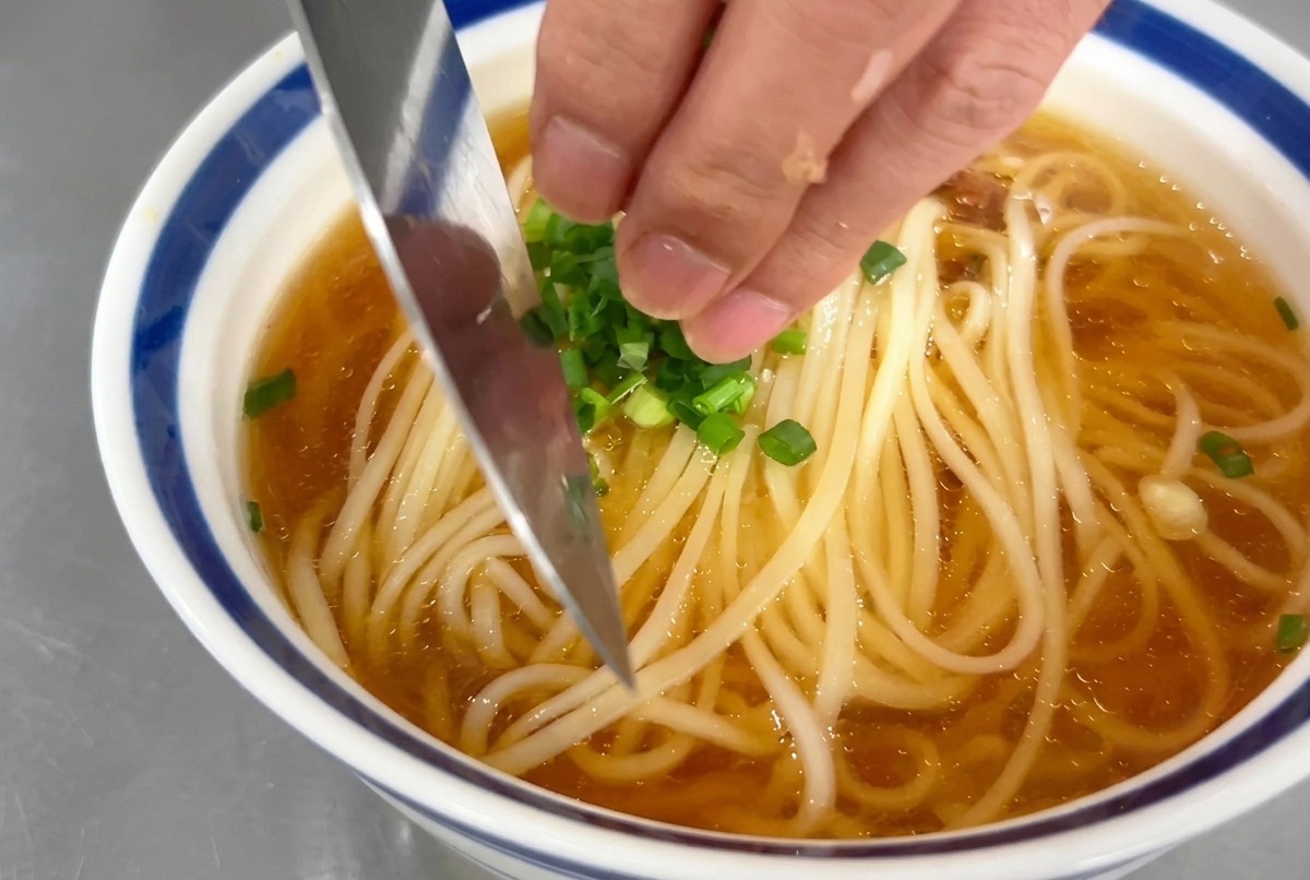 Una mano espolvoreando aros finos de cebolleta verde sobre el cuenco de sopa de fideos.