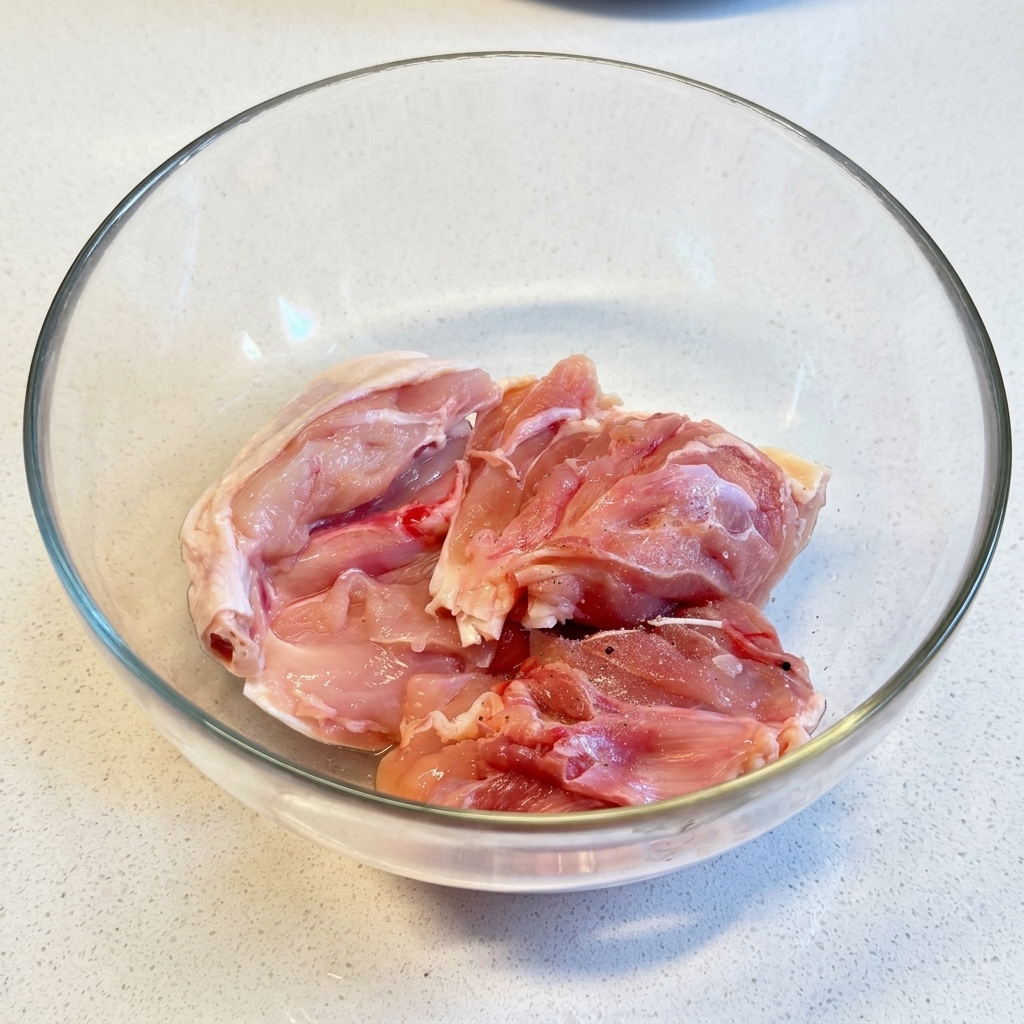 Contramuslos de pollo crudos sazonándose con sal y pimienta dentro de un tazón de vidrio transparente.