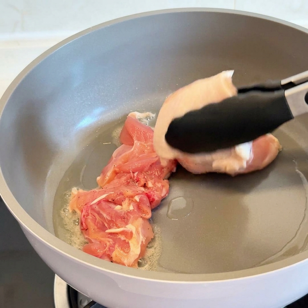 Unas pinzas colocando un contramislo de pollo crudo con la piel hacia abajo en una sartén antiadherente gris con aceite caliente.