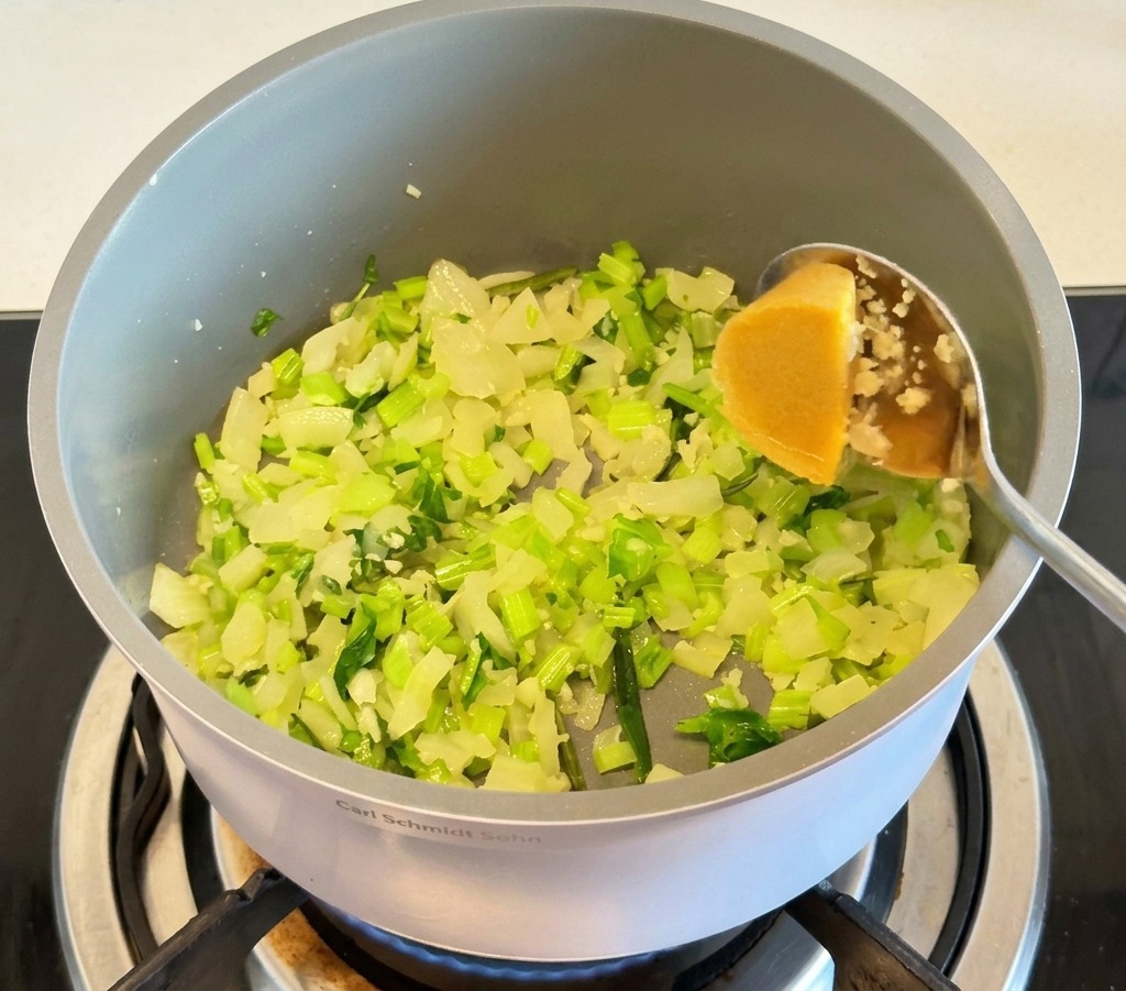 Una cuchara de metal añadiendo un cubo amarillo de caldo de pollo concentrado en una olla gris con trozos de cebolla y apio salteados.