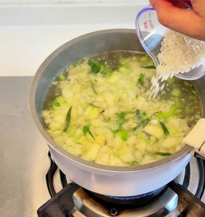 Arroz blanco vertiéndose desde una taza medidora transparente a una olla de sopa hirviendo con verduras y hierbas picadas visibles.