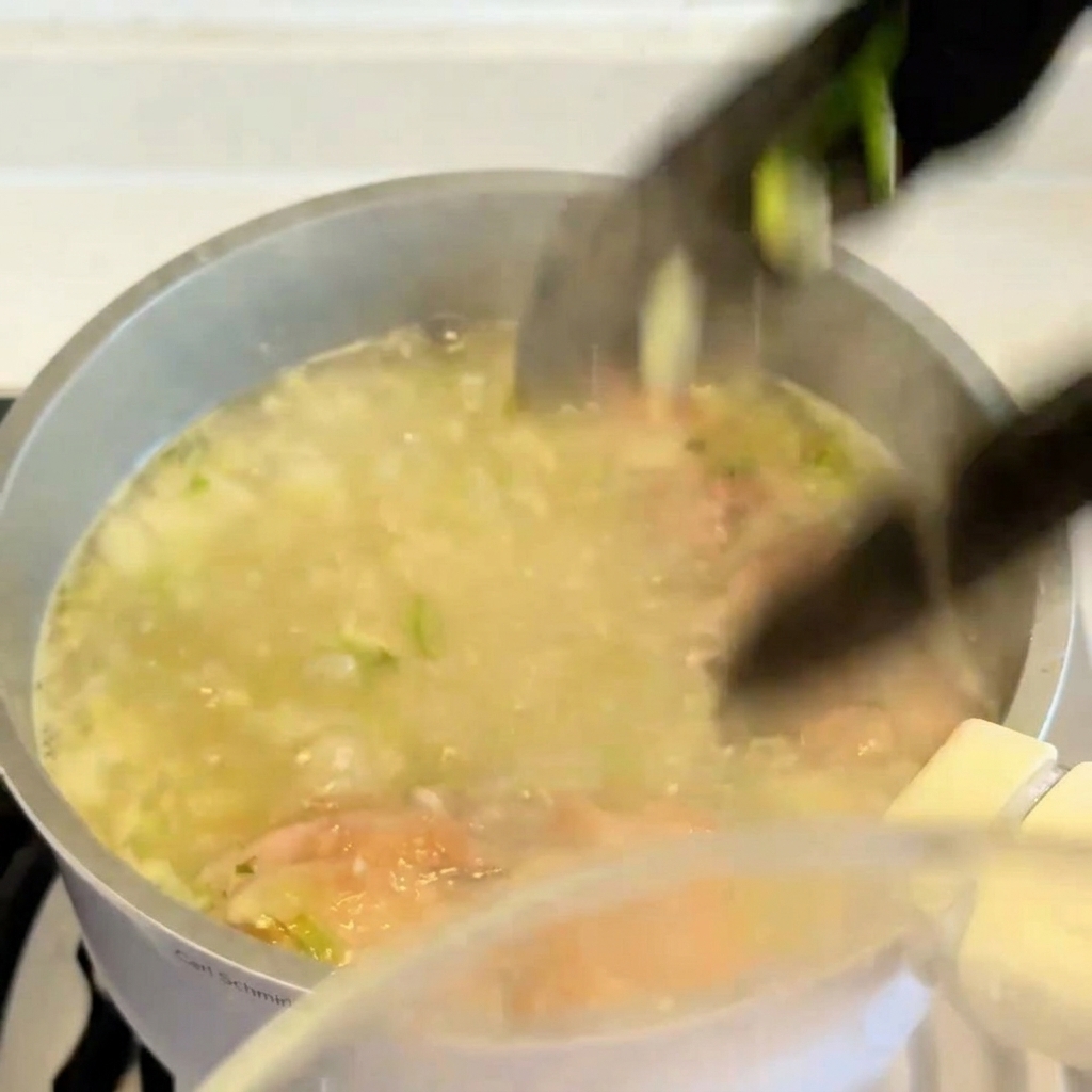 Un par de pinzas levantando un contramuslo de pollo cocido y dorado de una olla de caldo de sopa hirviendo.