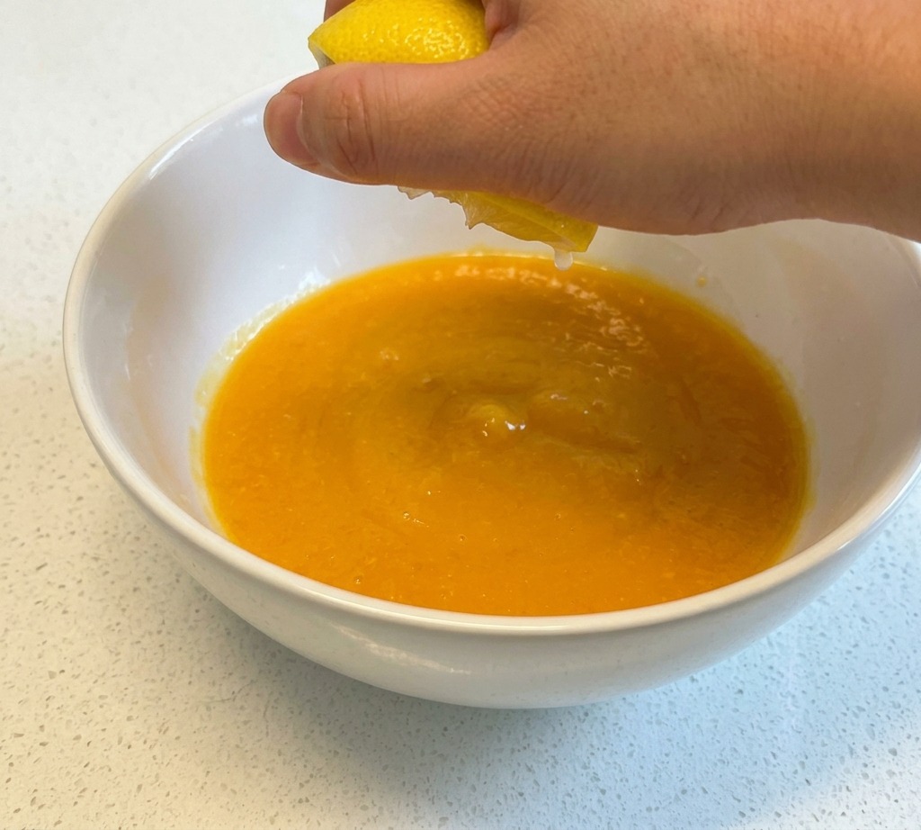 Una mano exprimiendo el jugo de medio limón amarillo en un tazón de cerámica blanca que contiene huevos batidos.