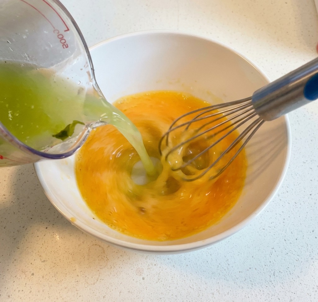 Caldo caliente vertiéndose desde una taza medidora a un tazón con huevos batidos y limón mientras se mezcla con un batidor de globo.