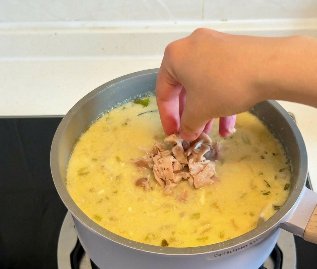 Manos dejando caer trozos de pollo cocido desmenuzado en una olla de sopa amarilla espesa y cremosa.