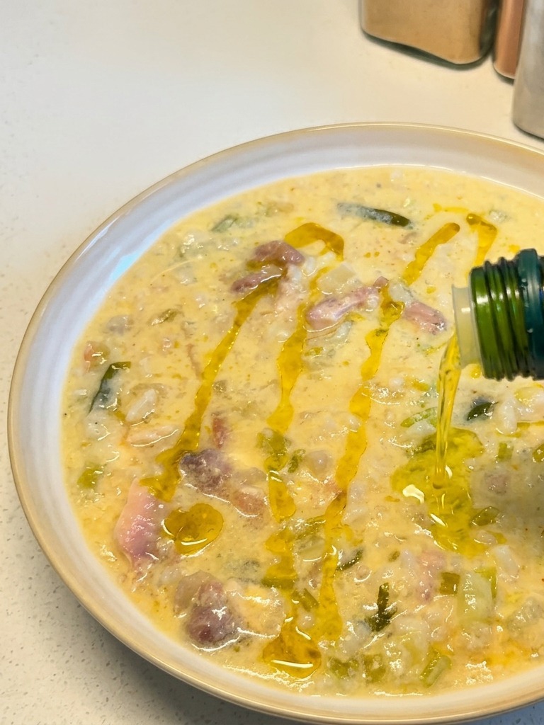 Aceite de oliva dorado vertiéndose desde una botella verde sobre un tazón servido de sopa cremosa de pollo y limón.