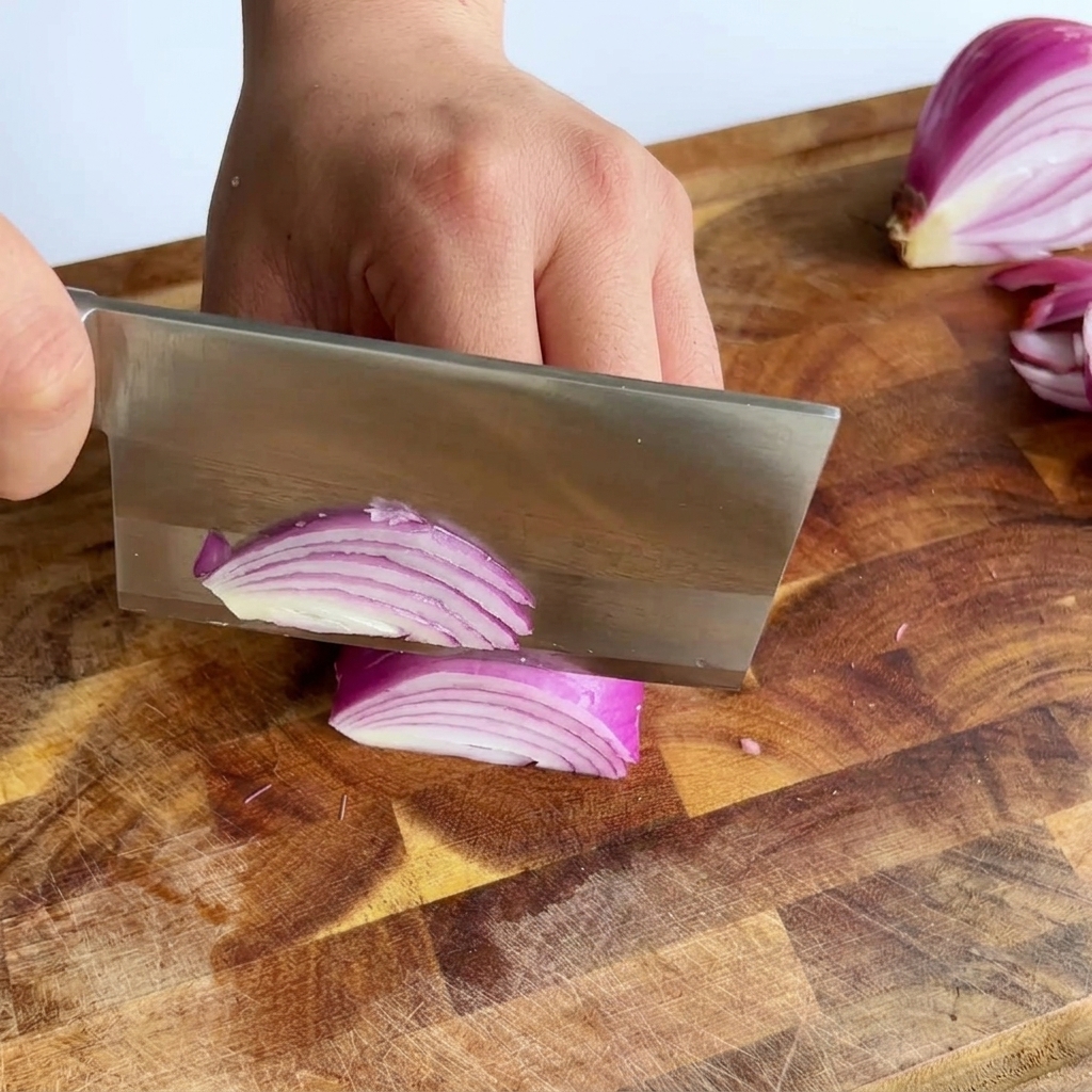 Un gran cuchillo cortando una cebolla morada para crear finas tiras púrpuras sobre una tabla de cortar de madera.