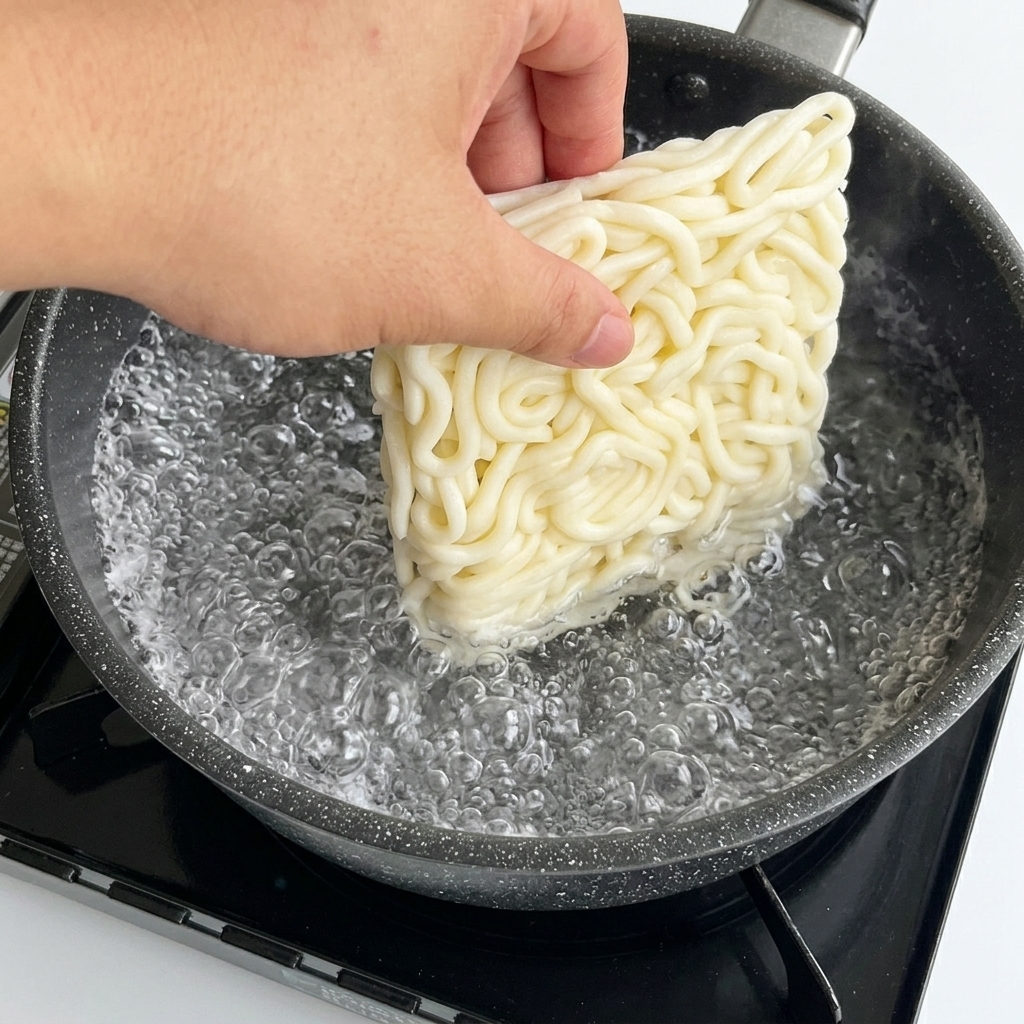 Una mano introduciendo un bloque cuadrado de fideos udon blancos en una olla de agua hirviendo.