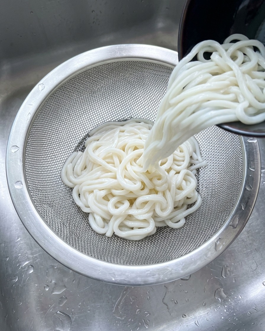 Fideos udon blancos siendo vertidos en un colador de malla de acero inoxidable bajo un chorro de agua fría en el fregadero.