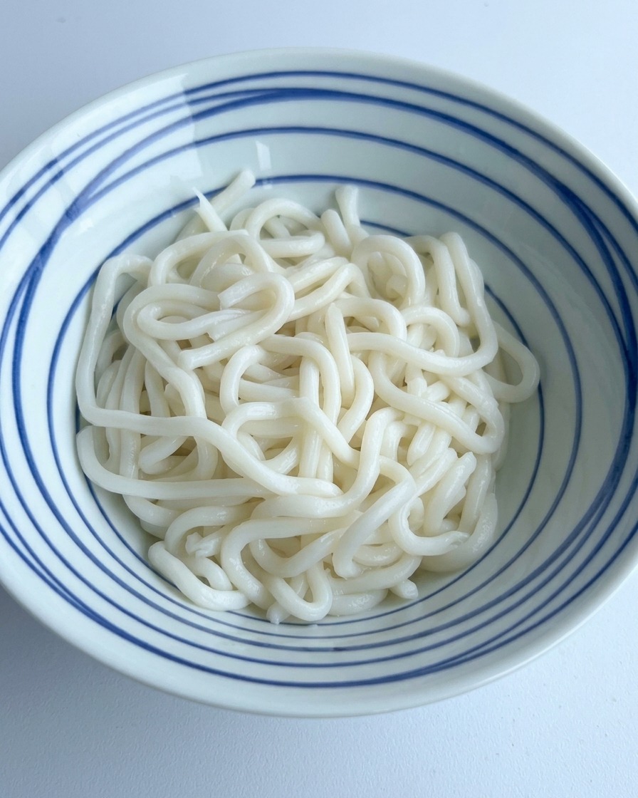 Salsa de soja oscura y condimentos siendo vertidos sobre una pila de fideos udon blancos en un tazón con rayas azules.
