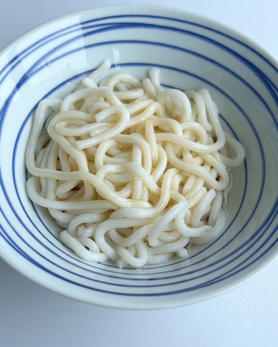 Agua clara siendo vertida en un tazón de fideos udon, mezclándose con los condimentos oscuros del fondo.