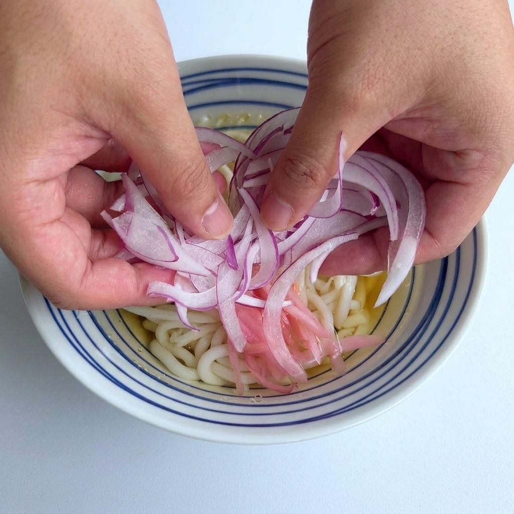 Dos manos dejando caer una porción de cebolla morada finamente picada en el centro de un tazón de fideos udon.