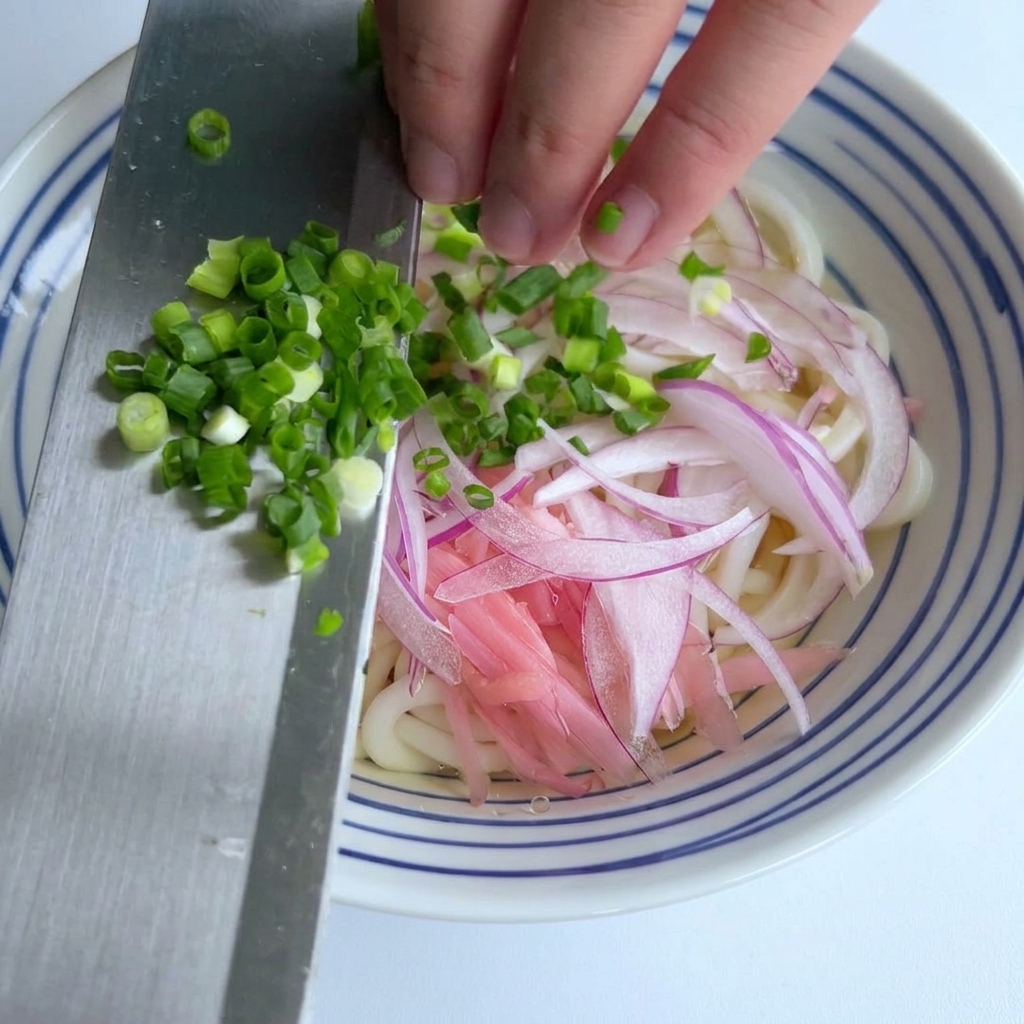 Cebollino finamente picado siendo deslizado desde el borde de un cuchillo metálico hacia un tazón de fideos udon con cebolla morada.