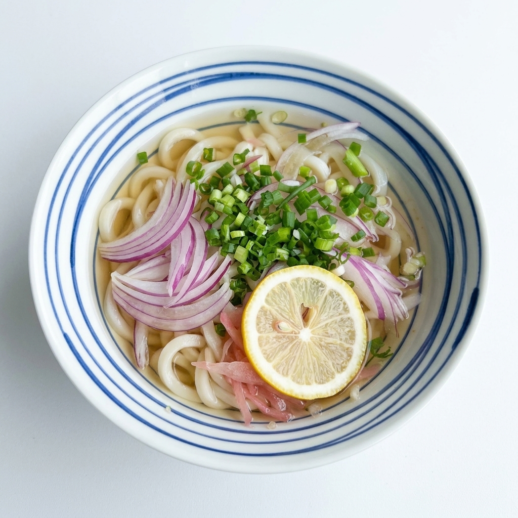 Vista aérea de un tazón de fideos udon en caldo claro, coronado con cebolla morada y cebollino, con una rodaja de limón amarillo en el centro.