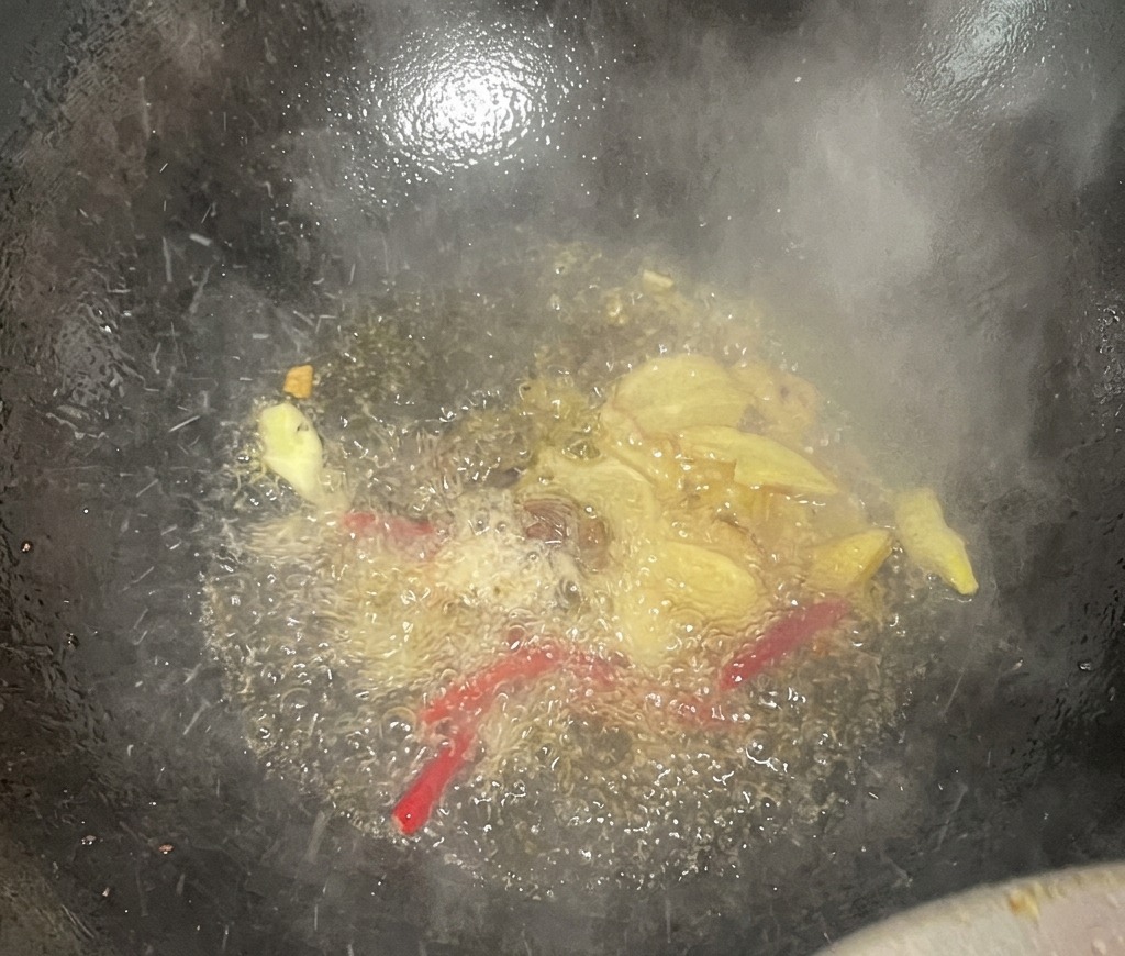 Rodajas de jengibre y chiles rojos secos burbujeando en aceite caliente dentro de un wok oscuro, soltando vapor y aromas.
