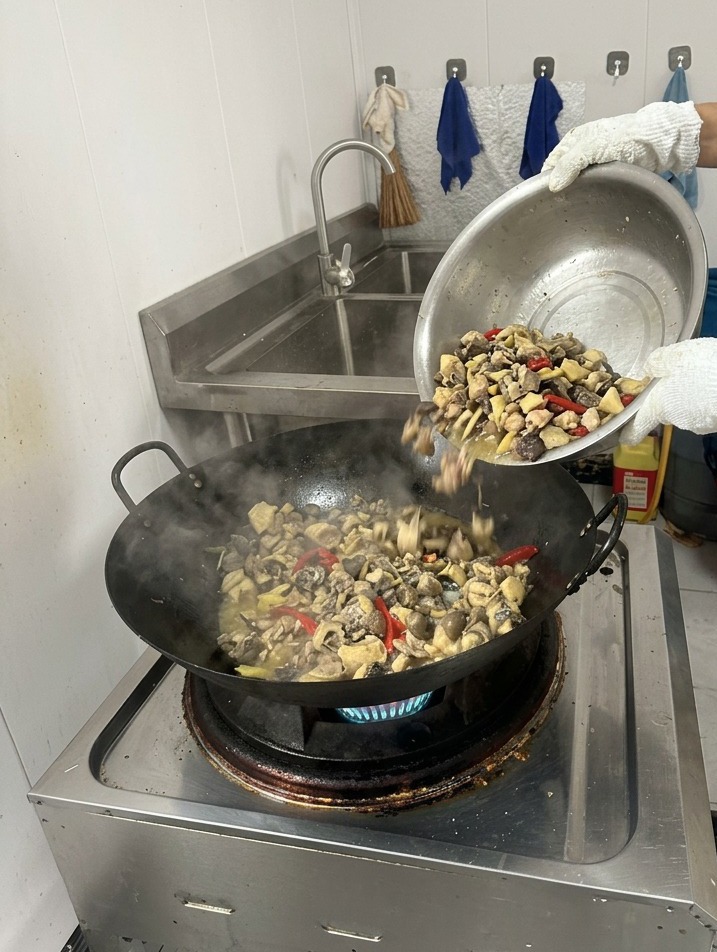 Una persona vertiendo agua hirviendo transparente desde una jarra hacia un wok humeante con pollo sellado y especias.