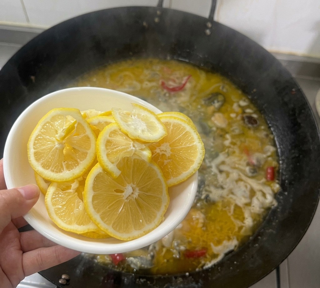 Una mano sosteniendo un cuenco blanco con rodajas finas de limón sobre un wok hirviendo lleno de caldo de pollo dorado.