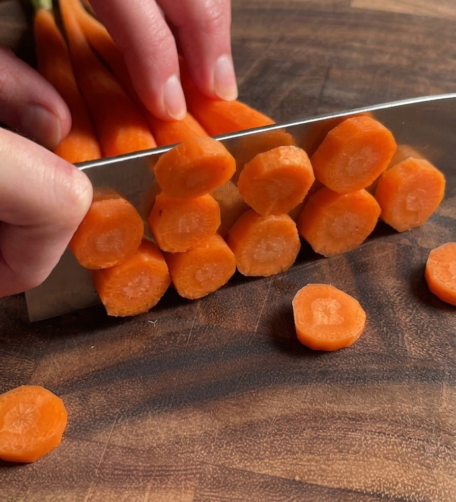Primer plano de unas manos usando un cuchillo para cortar varias zanahorias en rodajas finas sobre una tabla de cortar de madera oscura.