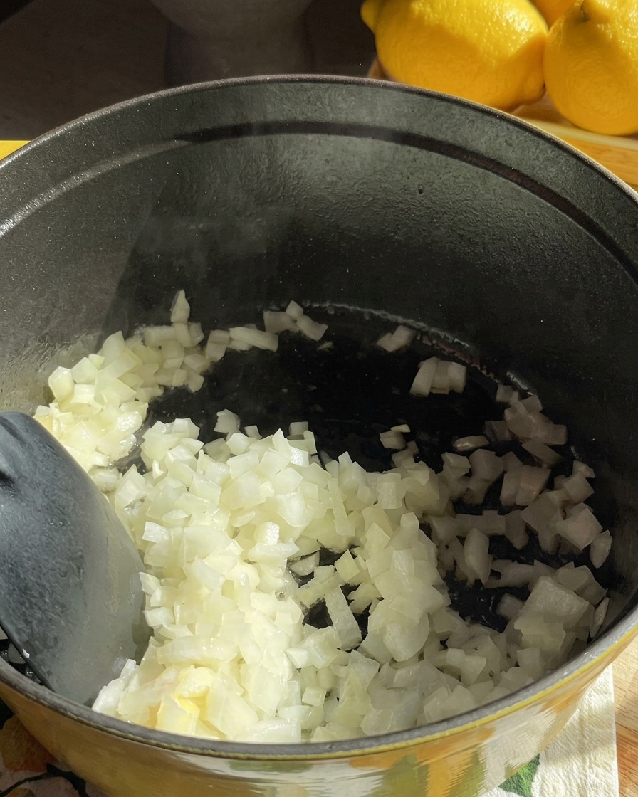 Cebolla blanca picada se remueve en una olla oscura mientras empieza a ablandarse en mantequilla.