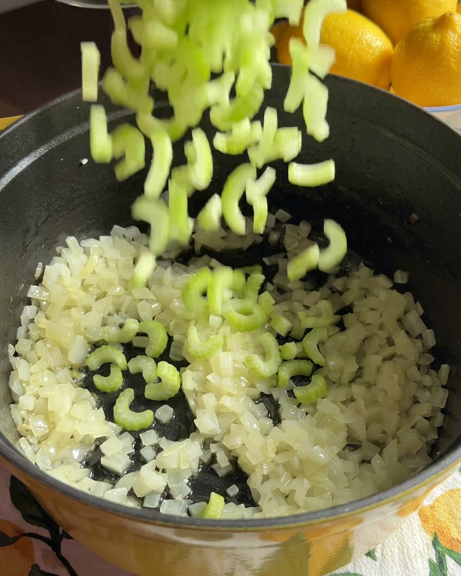 Trozos de apio verde picado caen en una olla que contiene cebolla y rodajas de zanahoria.